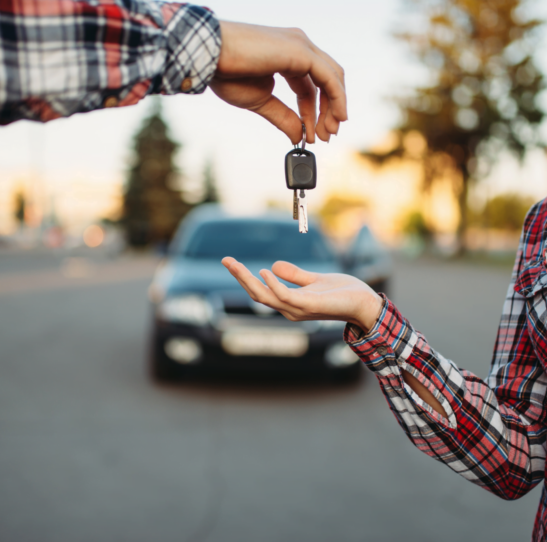 A person hands over car keys to a smiling young woman in a parking lot during the evening after her successful completion of Illinois Driver's Ed.